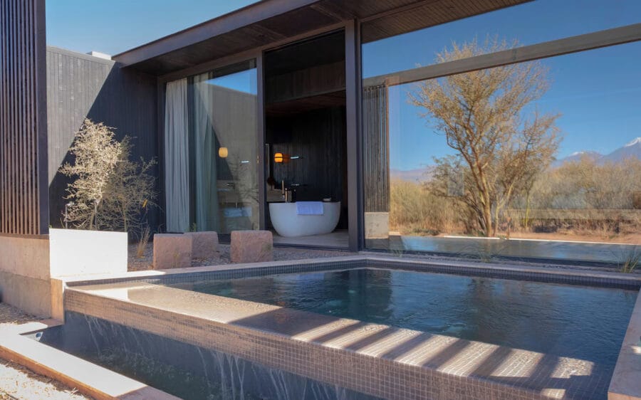 Modern building with private pool and large windows. Minimalist design, desert landscape visible outside. Small tree nearby, bathtub inside through glass. Tranquil atmosphere.