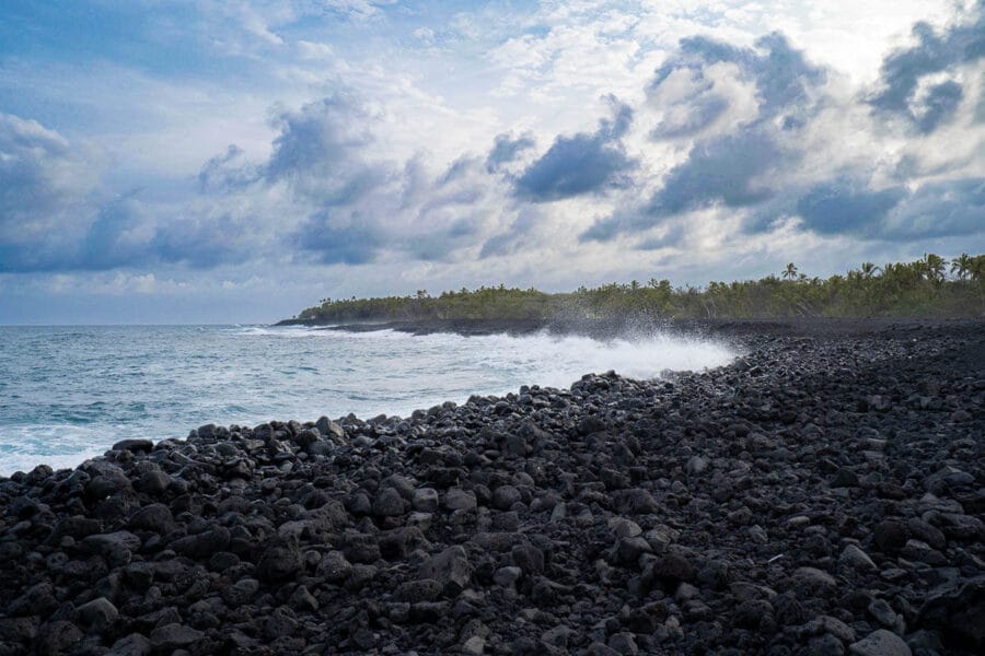 Rocky coastline with ocean waves crashing against black lava stones under a cloudy sky. Dense greenery lines the horizon, creating a dramatic scene.