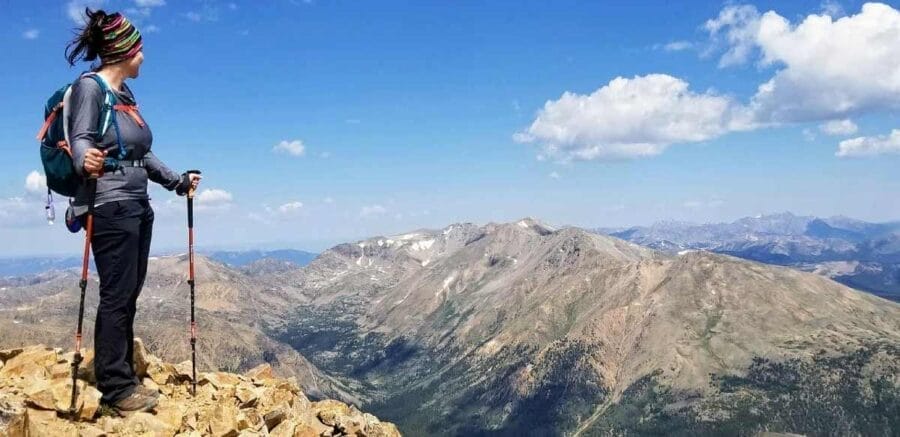 A person with hiking gear stands atop a mountain, admiring expansive views of the Rocky Mountains under a clear blue sky.