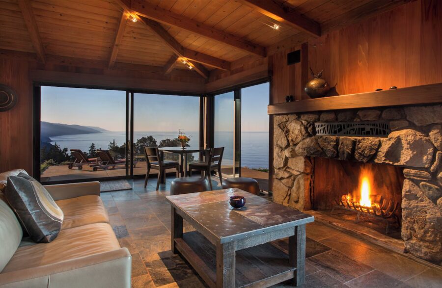 Cozy room with stone fireplace, wooden ceiling, and ocean view through large windows. A table with chairs offers a serene, inviting atmosphere.