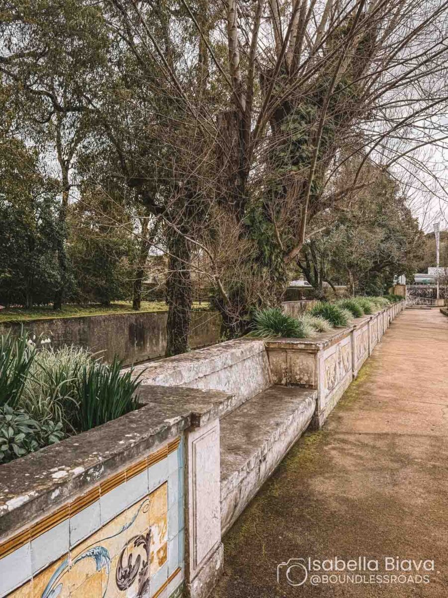 A scenic park path with decorated stone benches, surrounded by trees and greenery, and a person walking in the distance.