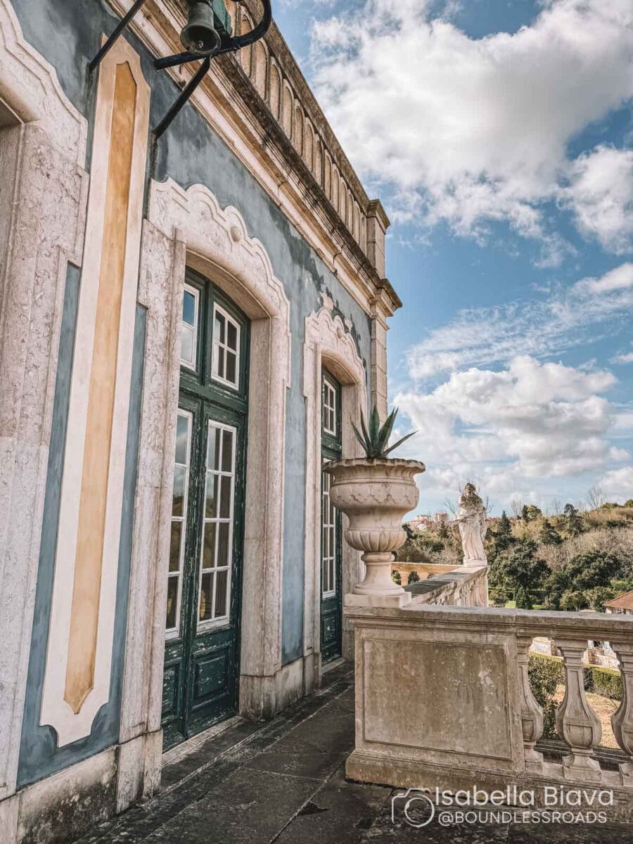 Historic building with ornate windows and balcony, featuring a decorative urn and statue under a partly cloudy sky. Scenic landscape visible in background.