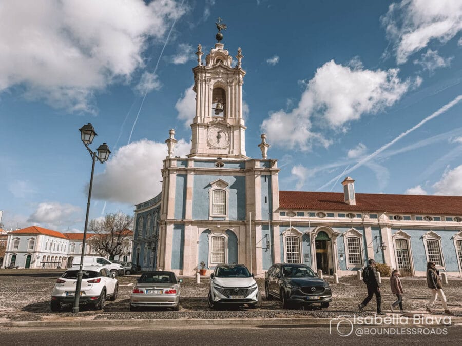 A grand historical building with a clock tower stands against a blue sky, with people and cars in the foreground.
