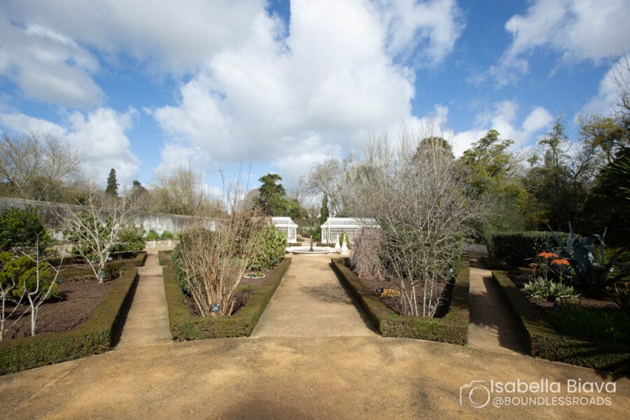 A formal garden with symmetrical pathways, bare winter trees, and hedges under a partly cloudy sky, featuring a central circular fountain.