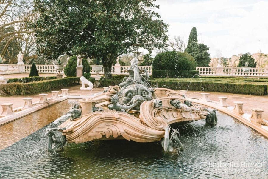 Ornate fountain with mythical figures and water spouts in landscaped garden, surrounded by statues and balustrades. Trees and bushes in the background.