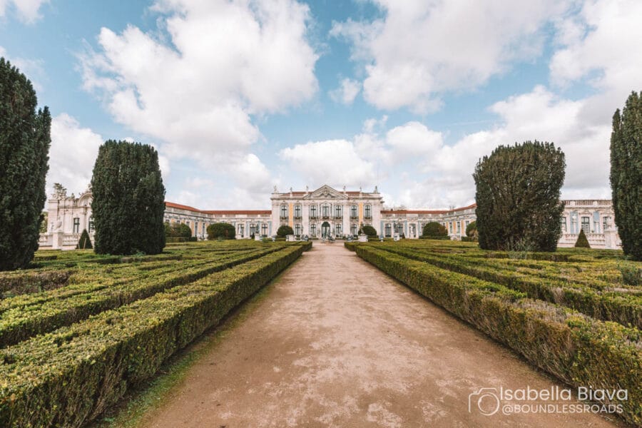 A grand palace with manicured gardens and pathways under a partly cloudy sky, framed by tall trees on both sides.