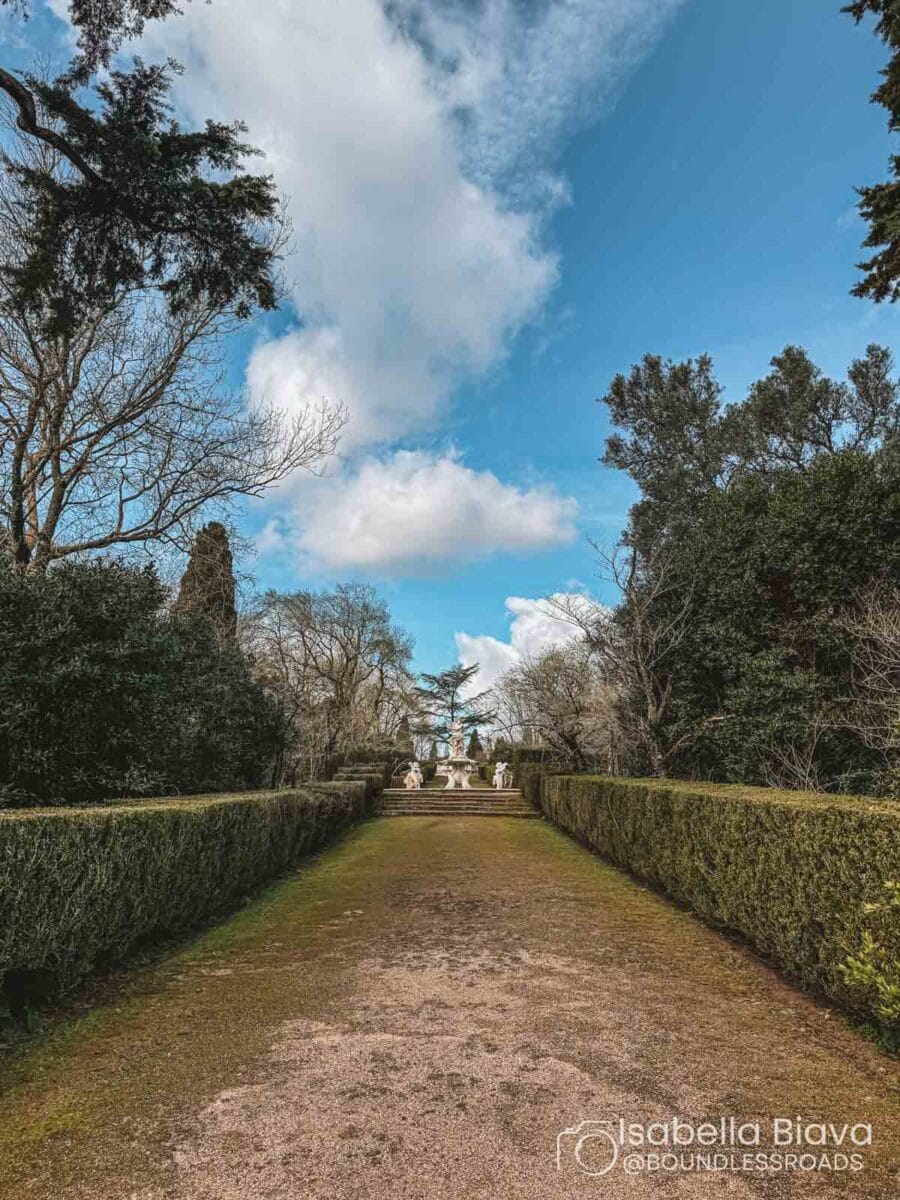 A tree-lined path leads to a statue garden under a bright, cloudy sky. Dense greenery surrounds the tranquil, scenic setting.