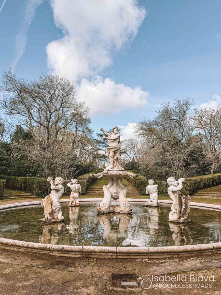 A Baroque-style fountain with intricate sculptures stands in a tranquil garden setting, surrounded by trees and hedges under a partly cloudy sky.