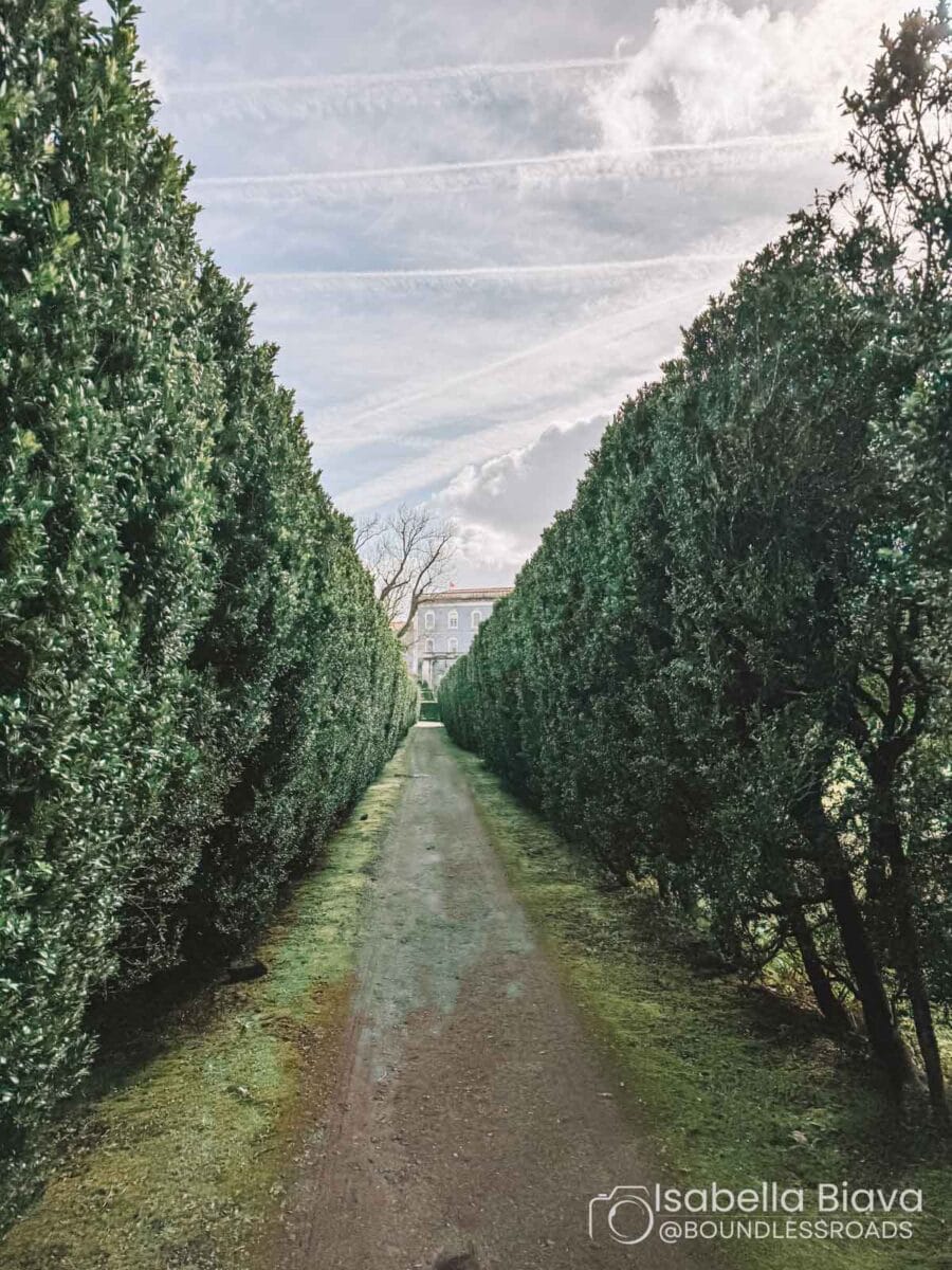 A narrow pathway flanked by tall hedges leads to a historical building in the distance, under a partly cloudy sky.