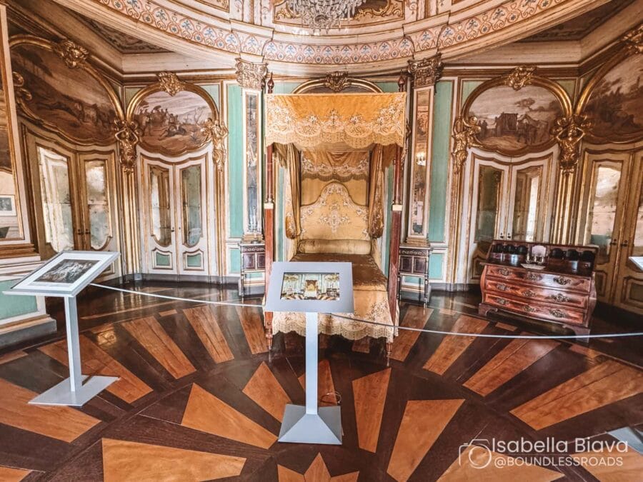 Ornate room in Royal Palace of Caserta with intricate decor, elegant canopy bed, informational displays, and wood-patterned floor reflecting opulent historical architecture.