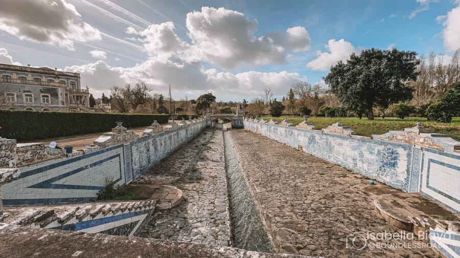 A long tiled canal stretches through the gardens of the Palace of Queluz, framed by lush greenery and a partly cloudy sky.