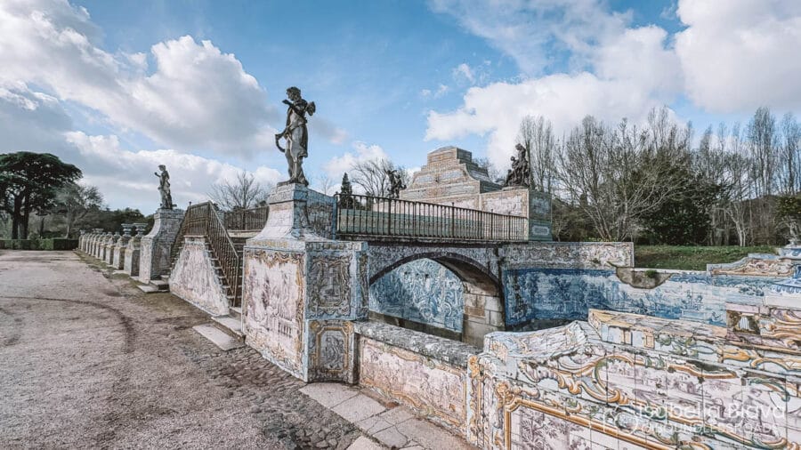 The image features the ornate azulejo tilework of the Palace of Queluz gardens, under a blue sky with scattered clouds and surrounding trees.