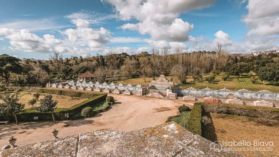 The image depicts the gardens of Quinta da Regaleira in Sintra, Portugal, featuring pathways and greenery under a partly cloudy sky.