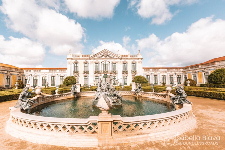A grand palace with intricate statues surrounds a central fountain under a bright sky, showcasing classic architecture and lush garden landscaping.