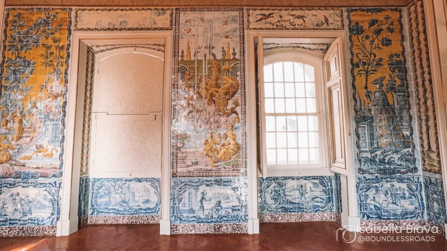 The image shows a room with intricate blue and yellow azulejo tiles, depicting various scenes, and featuring two arched windows.