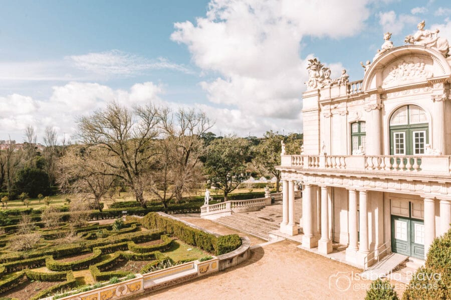 An elegant classical building with ornate details overlooks a manicured garden. The sky is partly cloudy, creating a serene, picturesque setting.