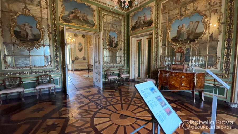The image shows an ornate, richly decorated room with intricate ceiling art and wooden flooring, featuring elegant furniture and information displays.