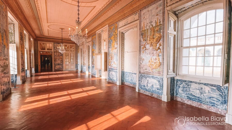 Ornate hallway with tiled walls, chandeliers, and arched windows. Sunlight streams through, creating patterns on the floor. Historical and elegant architectural details abound.