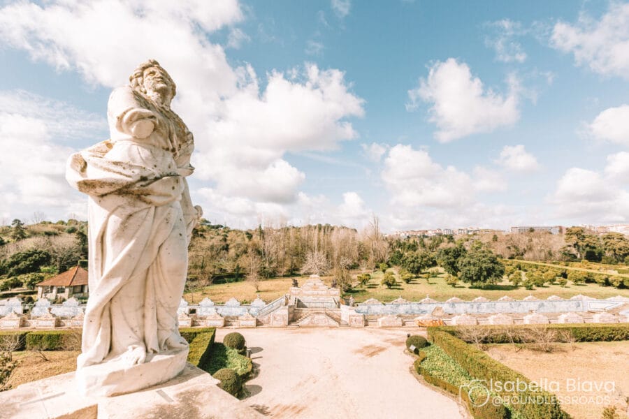 Stone statue overlooking Lisbon's Jardim Botânico Tropical with expansive gardens and lush trees under a bright, partly cloudy sky.