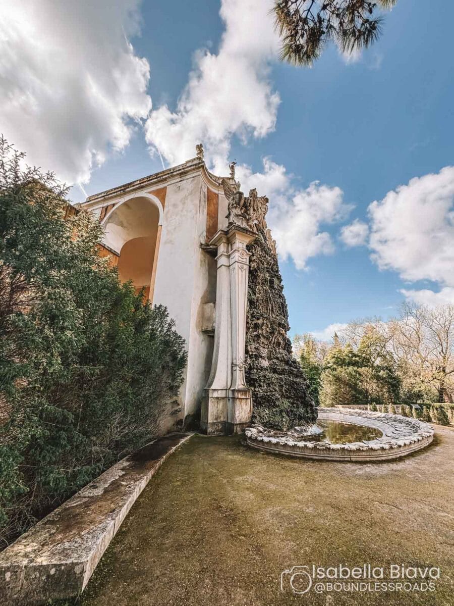 An ornate historic building with a fountain is set amid lush greenery under a partly cloudy sky, creating a serene atmosphere.