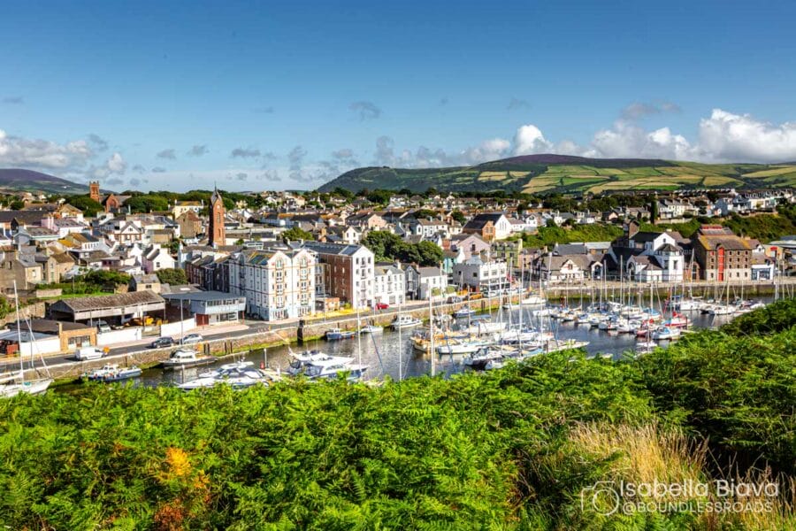 A picturesque view of Peel, Isle of Man, showcasing a marina with sailboats, historic buildings, rolling hills, and a quaint coastal town atmosphere.