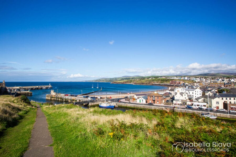 Coastal landscape with a path overlooking a town and harbor, clear blue sky, and hilly background. No recognizable landmarks or historical buildings visible.