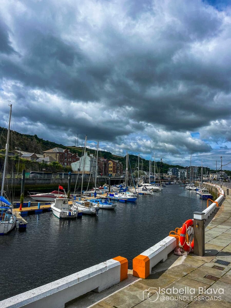 Cloudy sky over a marina with docked sailboats, surrounded by buildings and a walkway. The scene captures a tranquil and scenic harbor view.