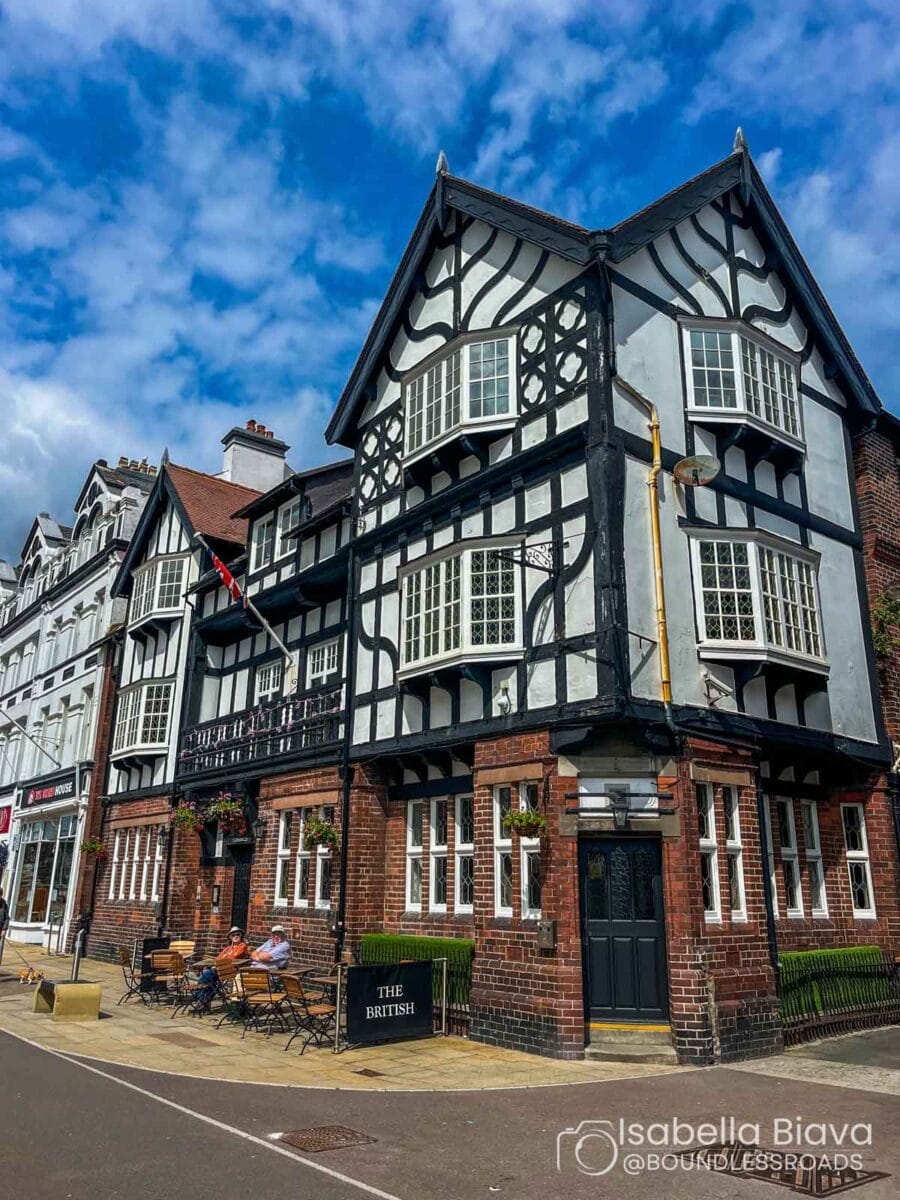Tudor-style building with black and white timber, "The British" pub sign. Several people seated outside. Traditional architecture, partly cloudy sky overhead.