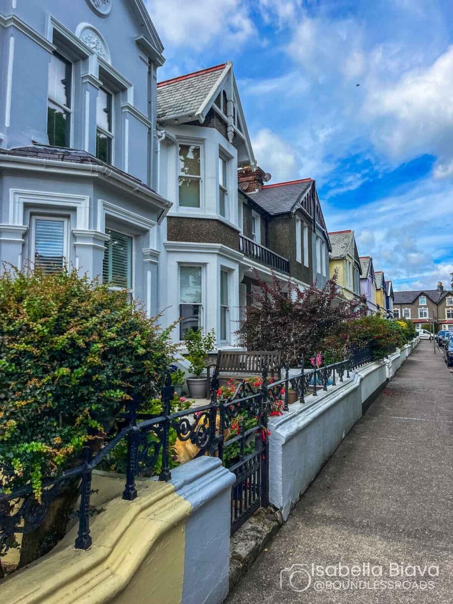 A row of Victorian-style terraced houses lines a quiet, tree-lined street on a cloudy day, featuring ornate iron railings and vibrant gardens.