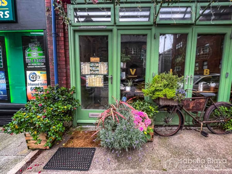 A quaint shopfront with green doors, bicycles, and lush plants outside. Displays vibrant flowers, creating a charming atmosphere. Vintage vibe on urban street.