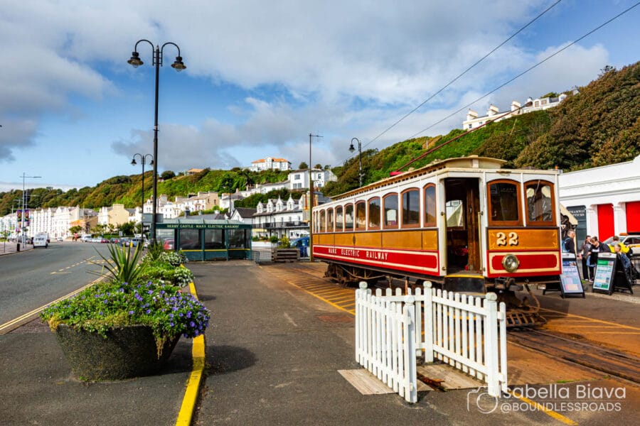 A vintage Manx Electric Railway tram is stationed near a promenade with coastal hills and white terraced buildings in the background, under a clear sky.