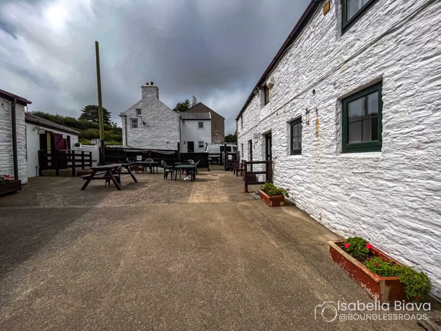 Courtyard with white stone buildings and outdoor seating. Overcast sky above. Potted plants line the walkway. Peaceful and rustic atmosphere.
