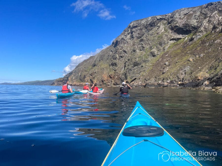 Three people kayaking in a calm, clear sea near rocky cliffs, under a blue sky with a few clouds.