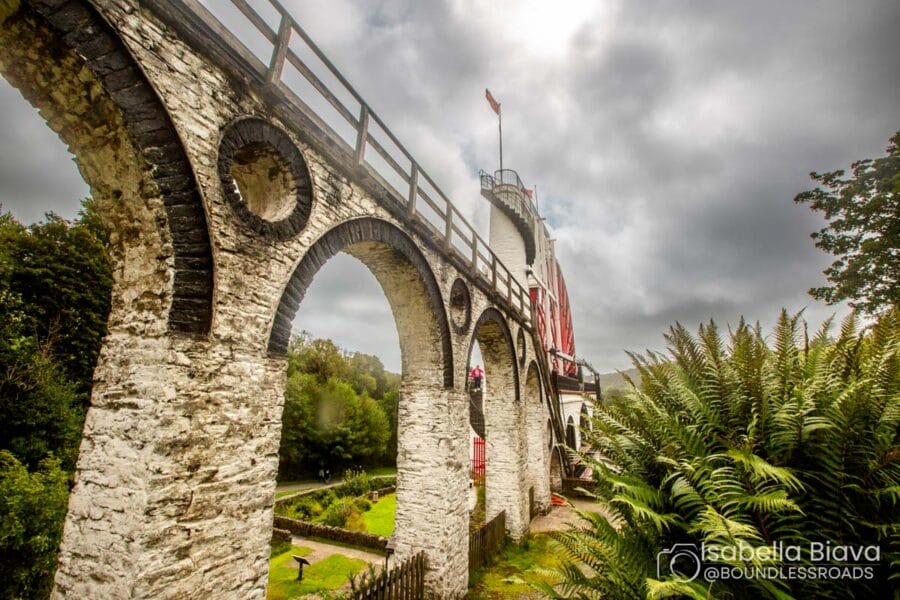 Stone arches of Devil's Bridge cross lush green landscape under cloudy skies, with a person standing nearby. Lush foliage frames the view.