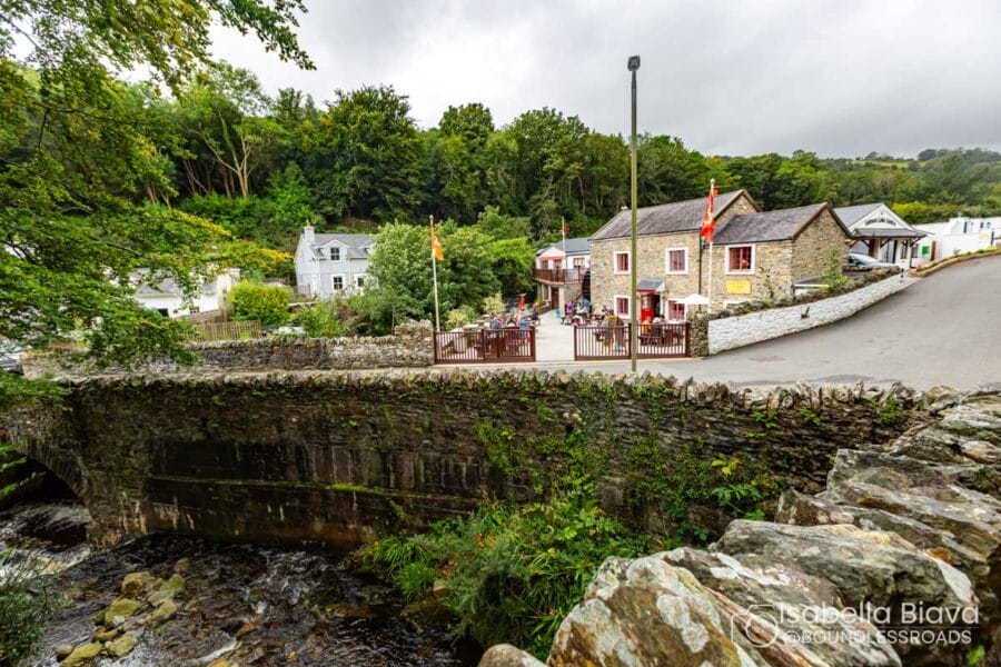 Stone bridge over a flowing stream, with charming stone buildings and people, surrounded by lush greenery. Historic architecture in a quaint village setting.