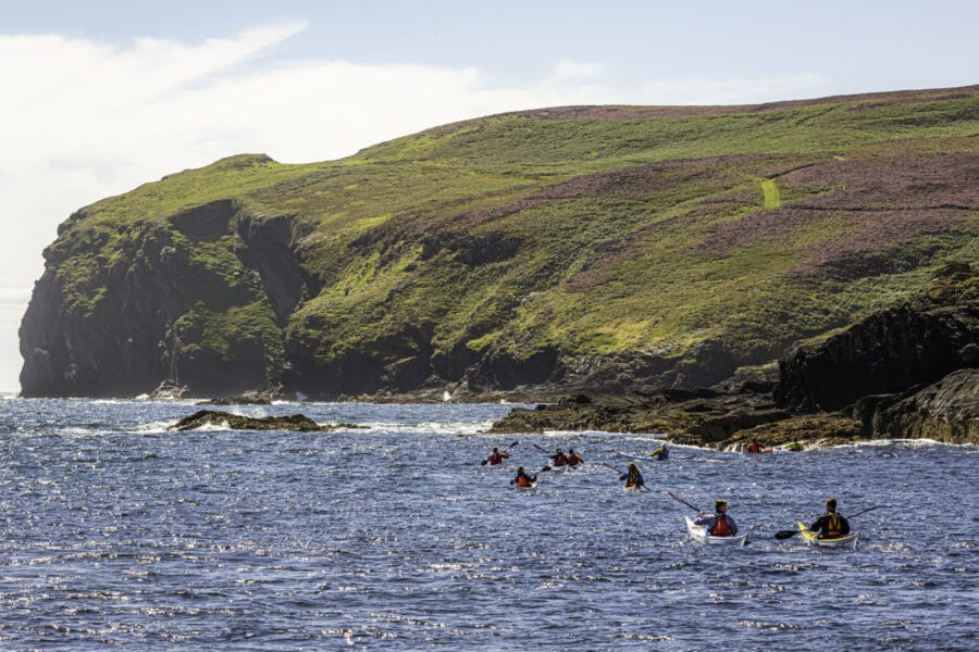 People kayaking near rocky, green cliffs under a partly cloudy sky. The scene suggests adventure and natural beauty along the coastline.