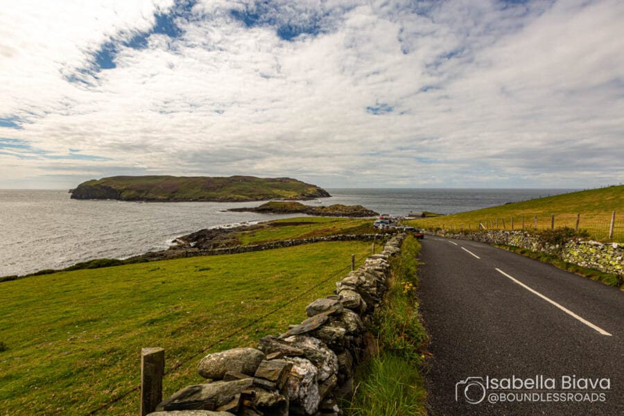 A coastal road on the Isle of Man, lined with stone walls, overlooks the sea and a grassy landscape under a partly cloudy sky.
