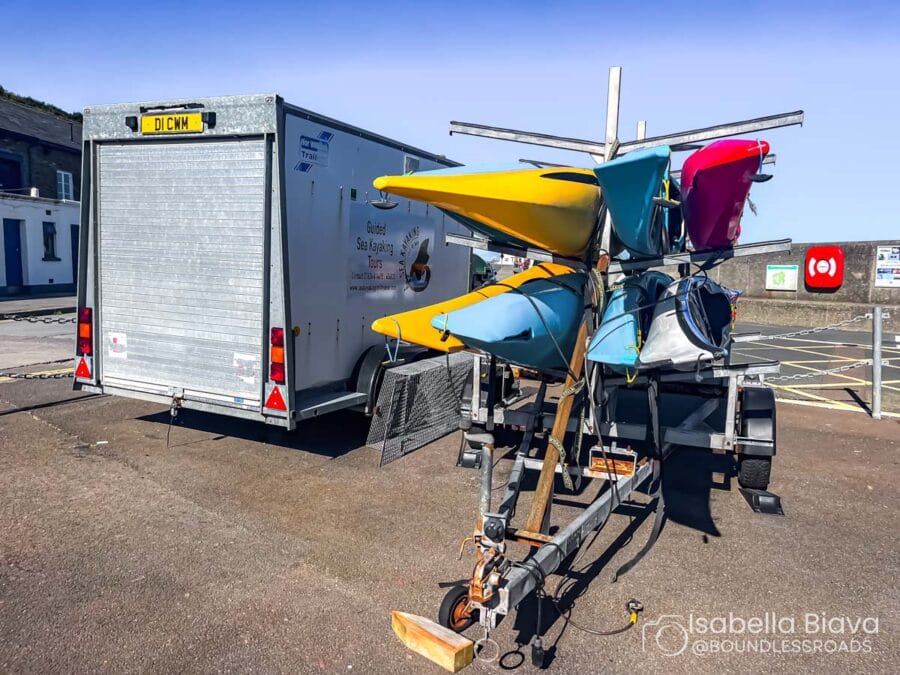 A trailer carries colorful kayaks next to a parked van in a coastal area. The clear sky suggests a sunny day.